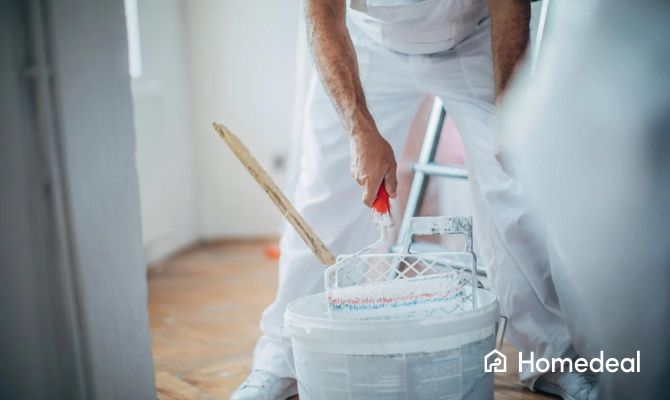 Schilder in witte werkkleding gebogen over een emmer met verf, terwijl hij een verfroller vasthoudt en gereedmaakt voor gebruik